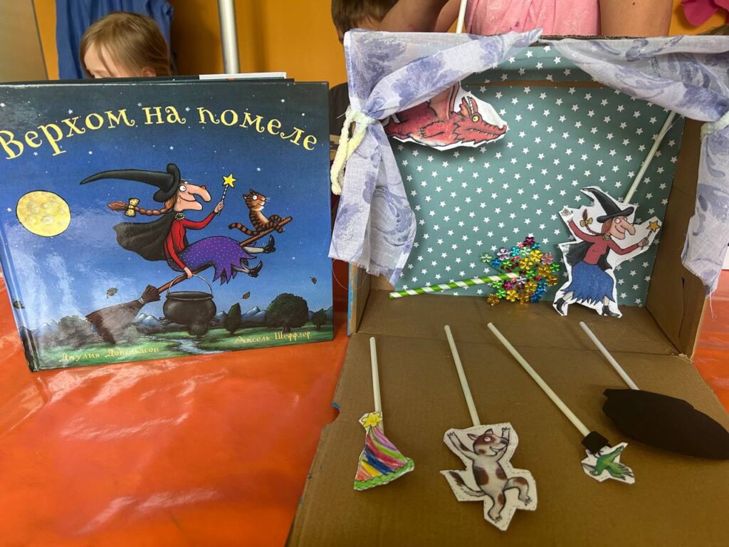 Ein Buch mit russischem Titel steht neben einer selbst gestalteten Spielfläche mit Papierfiguren an Stäben und weiteren Dekorationen.