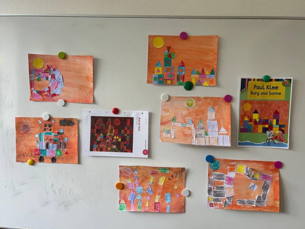 An einer Wand hängen mehrere farbenfrohe Kinderarbeiten mit geometrischen Formen neben einer Abbildung zu Paul Klees „Burg und Sonne“.
